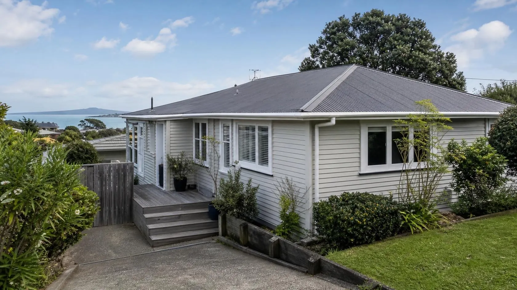 Single-storey pale grey weatherboard house with white window joinery, a dark hipped roof, white gutters and downpipe, a timber front deck with steps and a dark timber gate, beside a sloping concrete driveway with mature coastal shrubs, a large tree and distant sea and island views behind