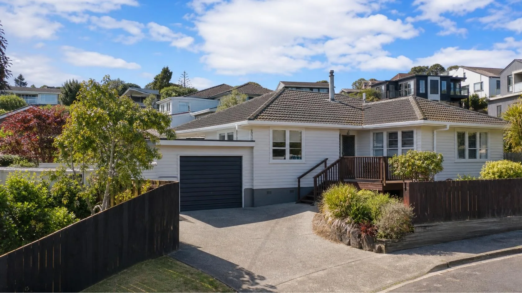 Single-storey white weatherboard house with a dark tiled hipped roof, grey roller garage door and a timber front deck with steps, beside a wide concrete driveway with dark timber fencing, mature trees and hillside homes behind