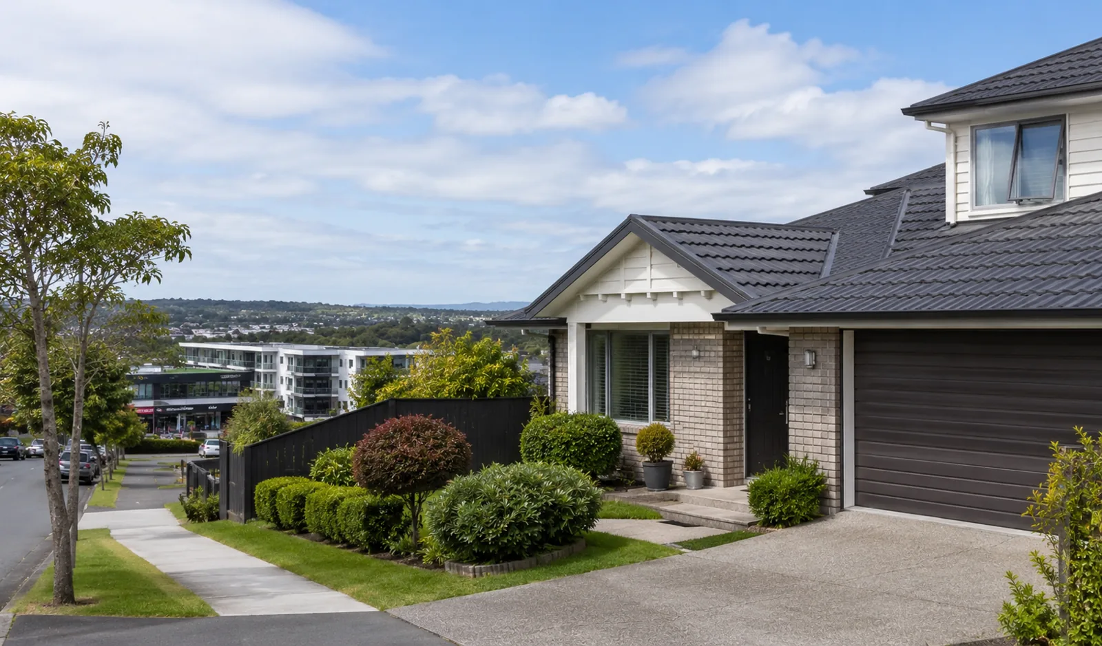 Two-storey light brick and white suburban home with dark garage, landscaped front garden and tree-covered hills in the distance