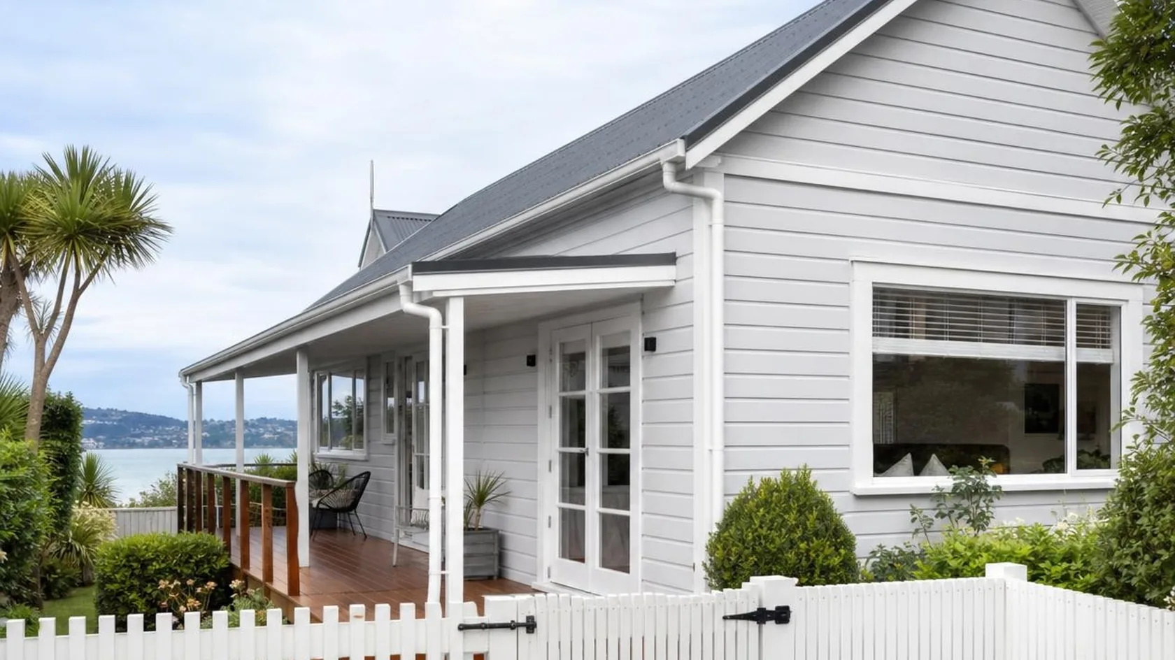 Single-storey pale grey weatherboard house with a dark gabled metal roof, white window joinery, white gutter and downpipe, a covered timber verandah with a stained timber deck and white French doors, and a white picket fence and gate on black hardware, with clipped hedges, a cabbage tree and a calm sea and distant coastline behind