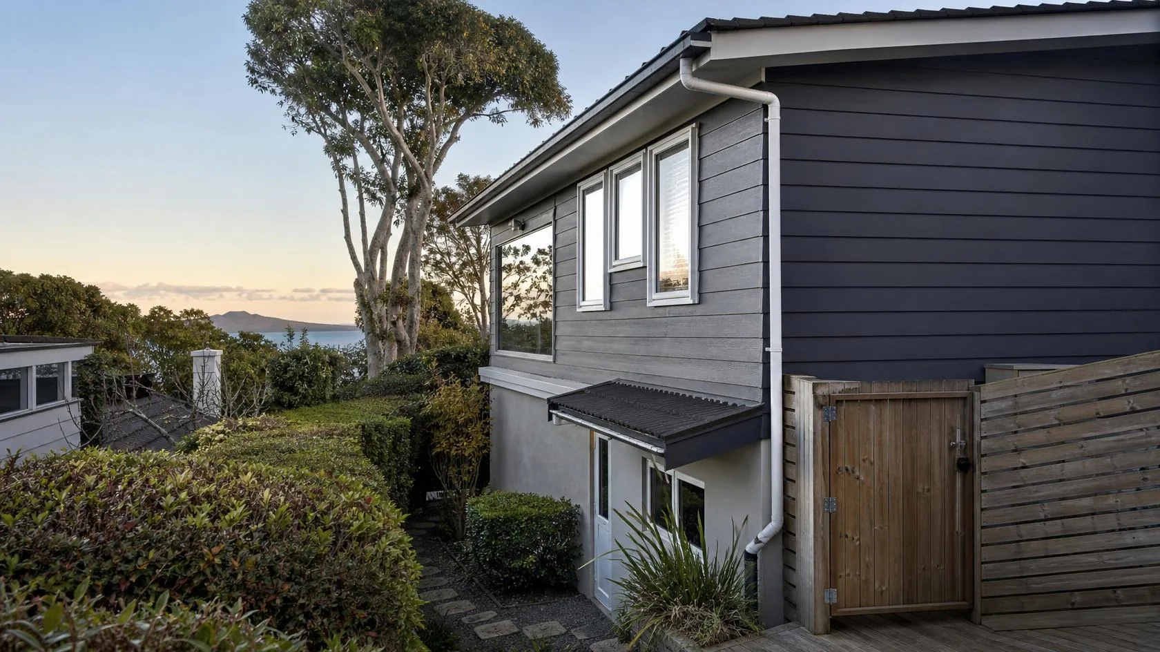 Two-storey home with grey and charcoal weatherboard cladding, white window joinery, a white gutter and downpipe, a small dark tiled awning over the lower entry door, and a timber gate in a slatted timber fence beside a deck, with clipped hedges, a large tree and a distant sea and island view behind