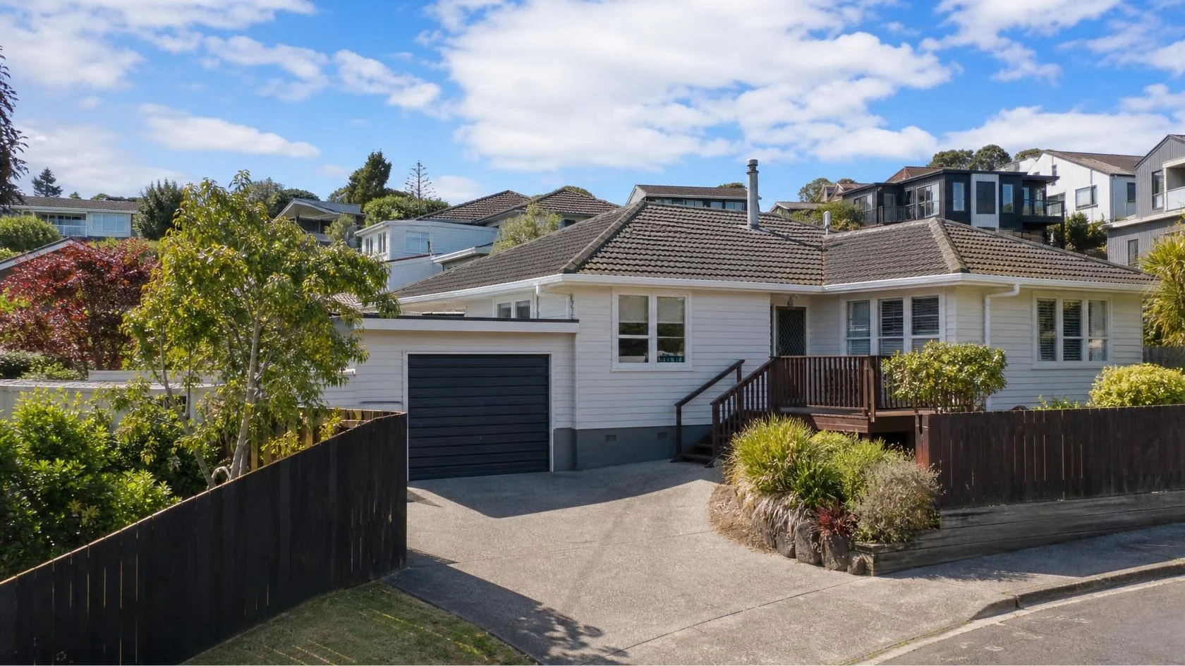 Single-storey white weatherboard house with a dark tiled roof, grey roller garage door and timber front deck beside a concrete driveway, with dark timber fencing and tree-covered hills behind