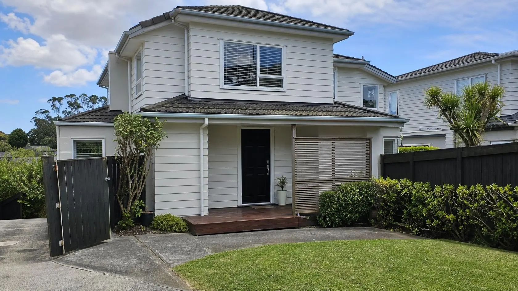 Two-storey cream weatherboard house with a dark tiled roof, black front door and timber entry deck, beside a concrete driveway with a dark timber gate, clipped hedge and lawn
