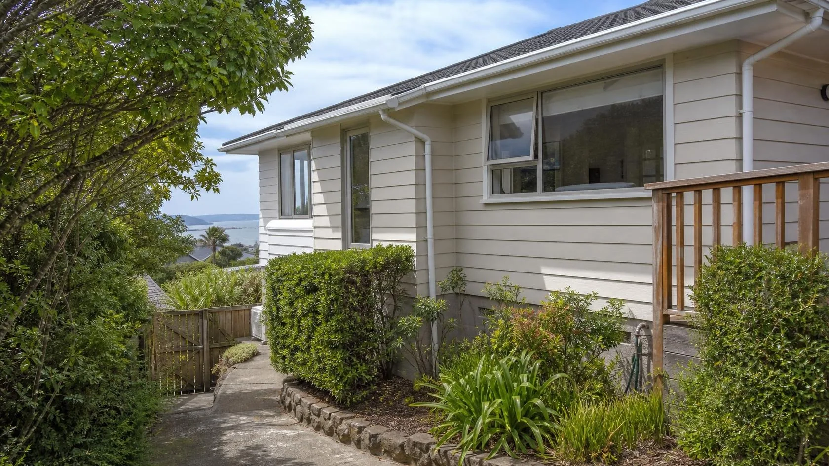 Single-storey pale grey weatherboard house with a dark tiled hipped roof, white window joinery, a white gutter and downpipe, a timber deck balustrade and a timber gate, with clipped hedges, a stone-edged garden border beside a concrete path, a large leafy tree and a calm sea and distant headland behind