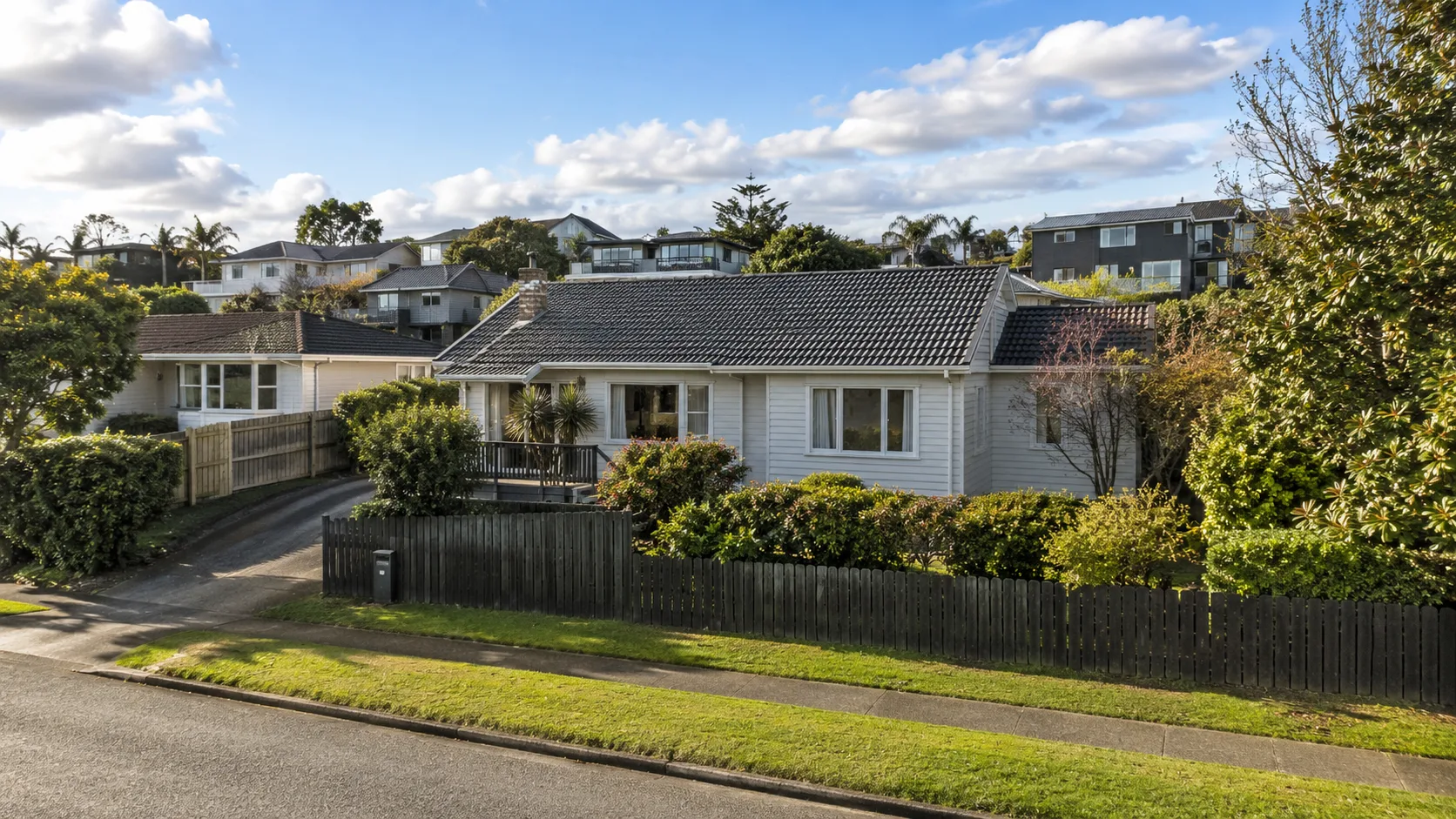 Single-storey off-white weatherboard house with a dark grey tiled gabled roof and a dark vertical-slat front fence, manicured lawn and hedges, on a hill with other homes and trees behind